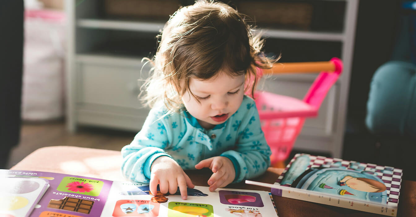 Kleinkind in blauem Schlafanzug betrachtet konzentriert ein buntes Bilderbuch im Spielzimmer mit Spielzeug im Hintergrund.