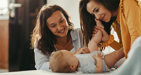 Zwei lächelnde Frauen spielen mit einem glücklichen Baby, das auf einem Bett liegt.