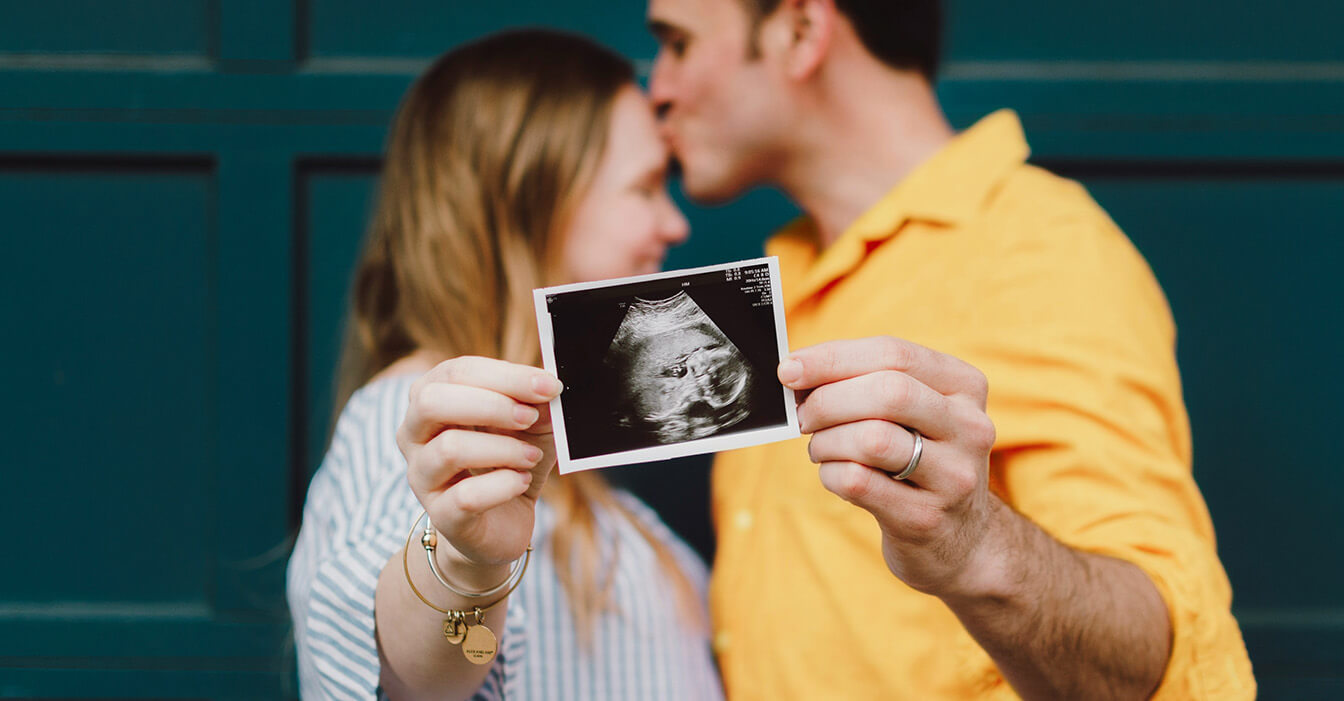 Paar hält Ultraschallbild eines Babys in die Kamera und küsst sich.